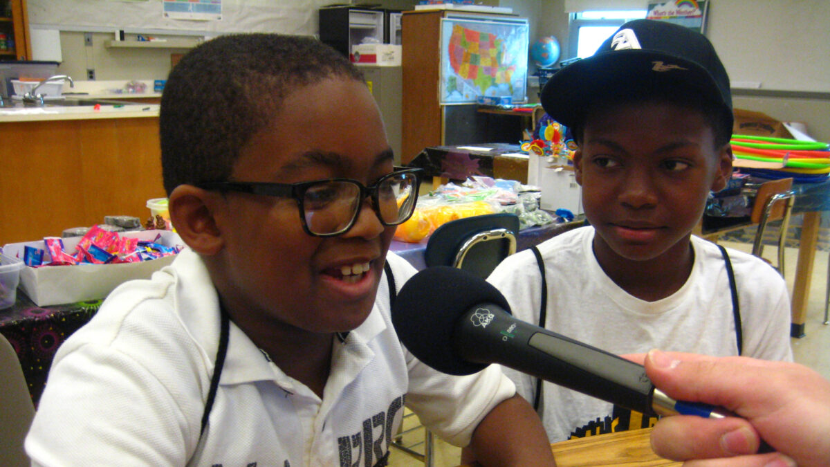 A youth looking at another youth speaking into a held microphone in a classroom