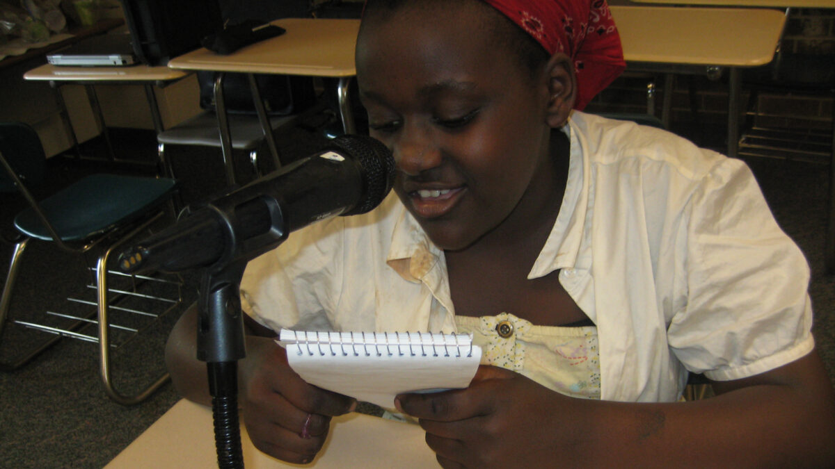 A youth holding a notepad and speaking into a microphone while sitting at a classroom desk