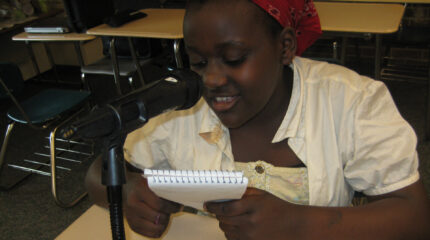 A youth holding a notepad and speaking into a microphone while sitting at a classroom desk