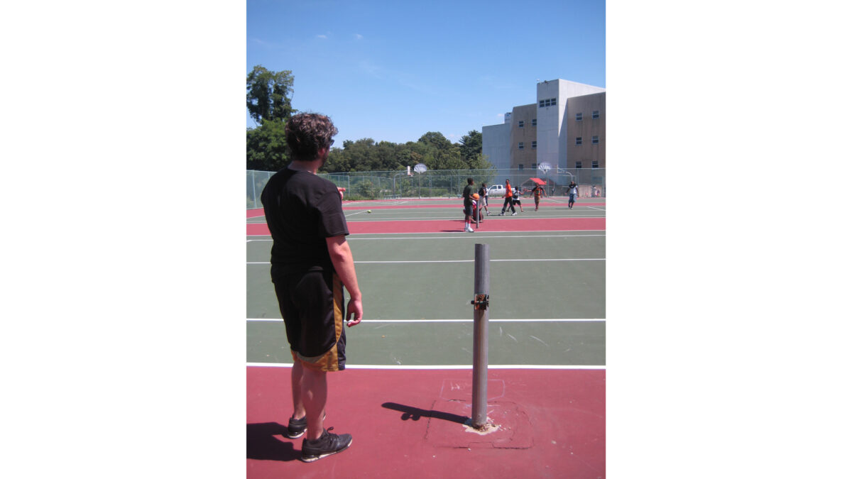 An adult standing next to a sawed-off metal pole watching youth playing with a kickball