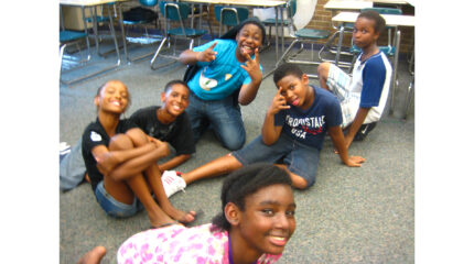 Six youth sitting on the floor smiling and posing for the camera in a classroom