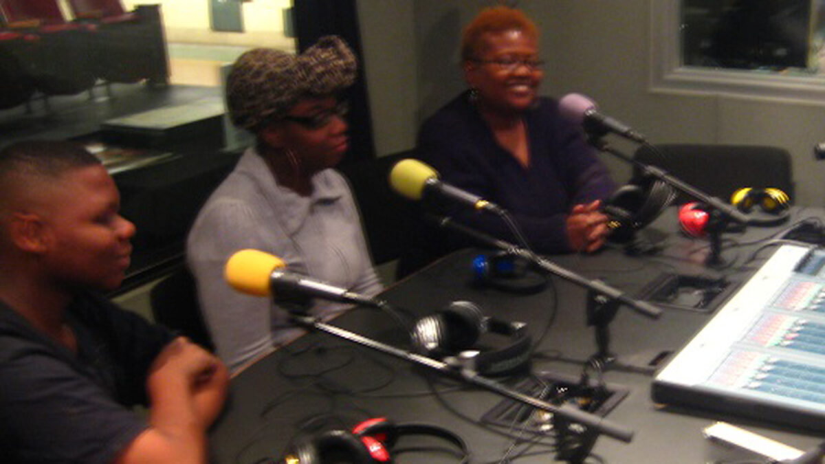 Three adults sitting behind microphones and headphones in a radio studio