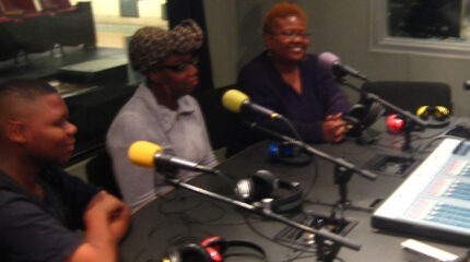 Three adults sitting behind microphones and headphones in a radio studio