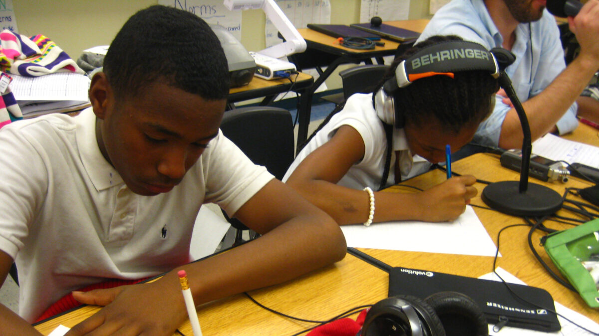Two youth, one wearing headphones, writing on pieces of paper on classroom desks