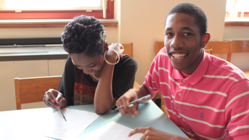 Two teens smiling and writing on pieces of paper in a library