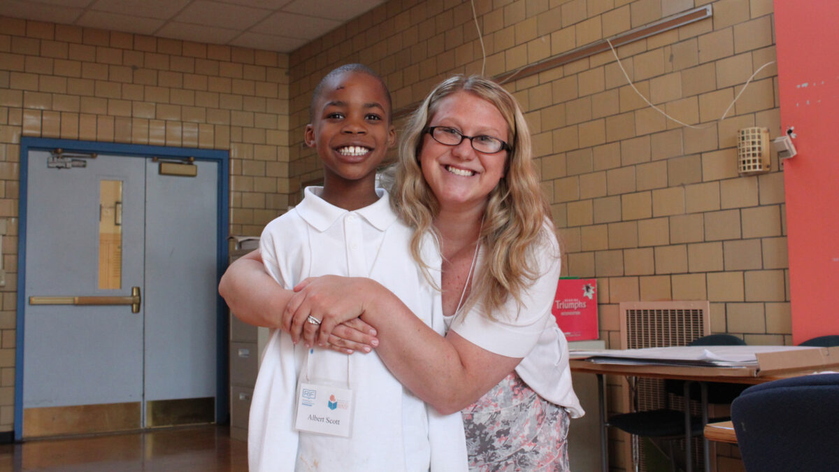 An adult wrapping their arms around a youth, both smiling at the camera in a classroom