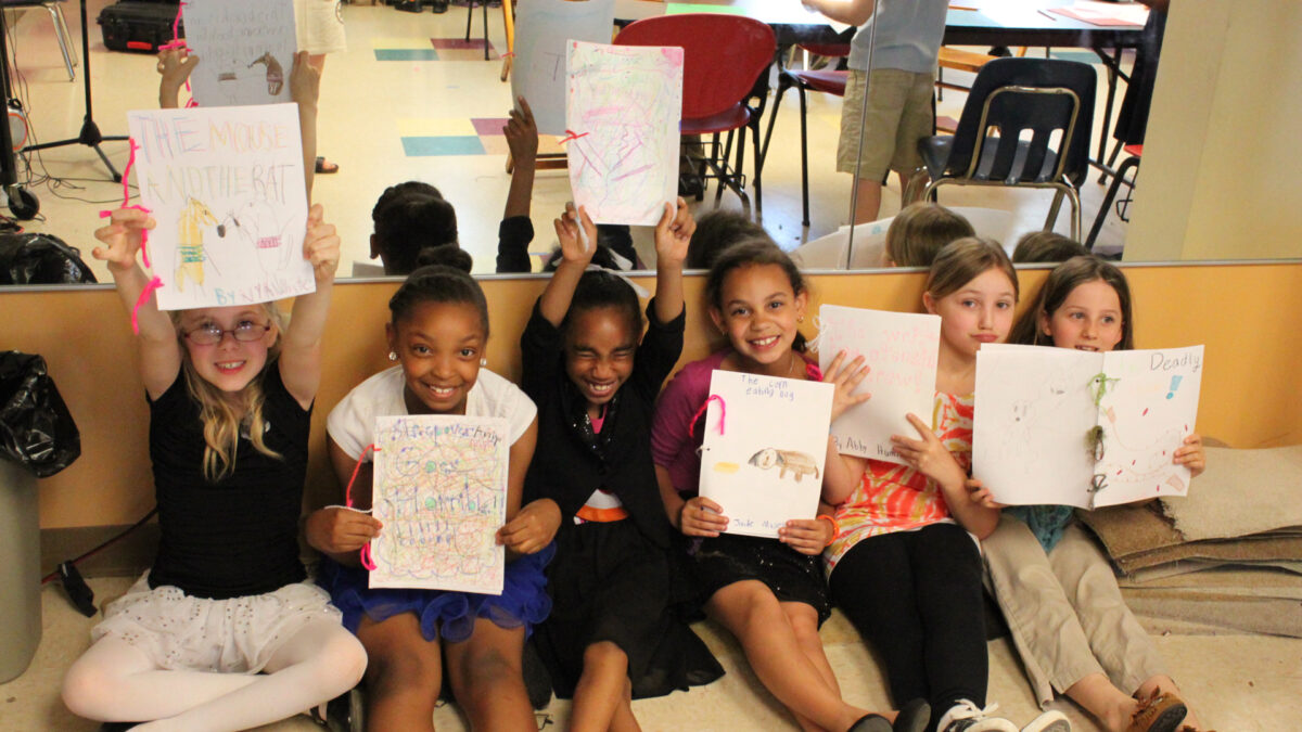 Six youth smiling and holding up drawings while sitting on the floor in front of a mirror