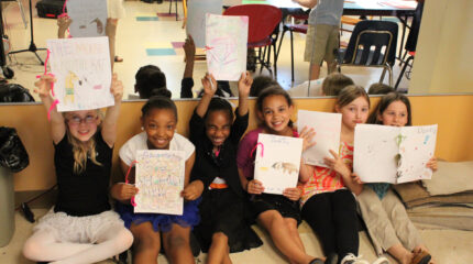 Six youth smiling and holding up drawings while sitting on the floor in front of a mirror