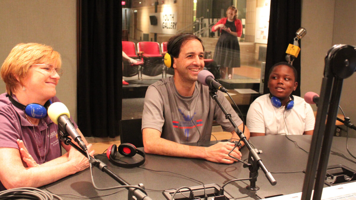 An adult wearing headphones around their neck, an adult wearing headphones on their head, and a youth wearing headphones around their neck, all sitting behind microphones in a radio studio