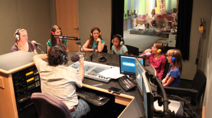Six youth wearing headphones and speaking into microphones being interviewed by an adult wearing headphones and speaking into a microphone sitting behind a mixing board in a radio studio
