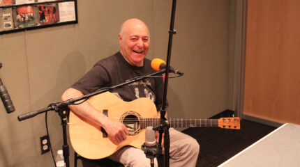 An adult smiling and sitting behind a microphone while holding an acoustic guitar in a radio studio