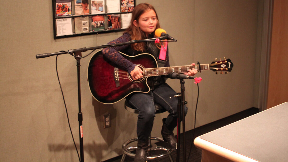 A youth sitting behind a microphone and holding an acoustic guitar in a radio studio