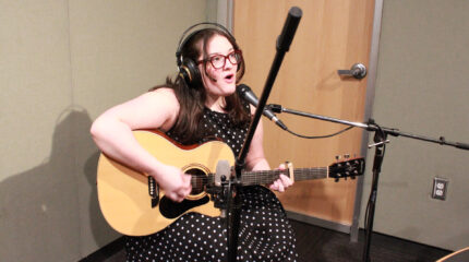 A teen playing an acoustic guitar and singing into a microphone in a radio studio