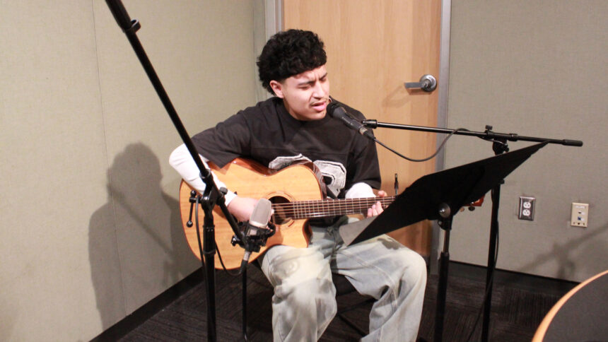 A teen sitting behind a music stand and playing an acoustic guitar while singing into a microphone in a radio studio