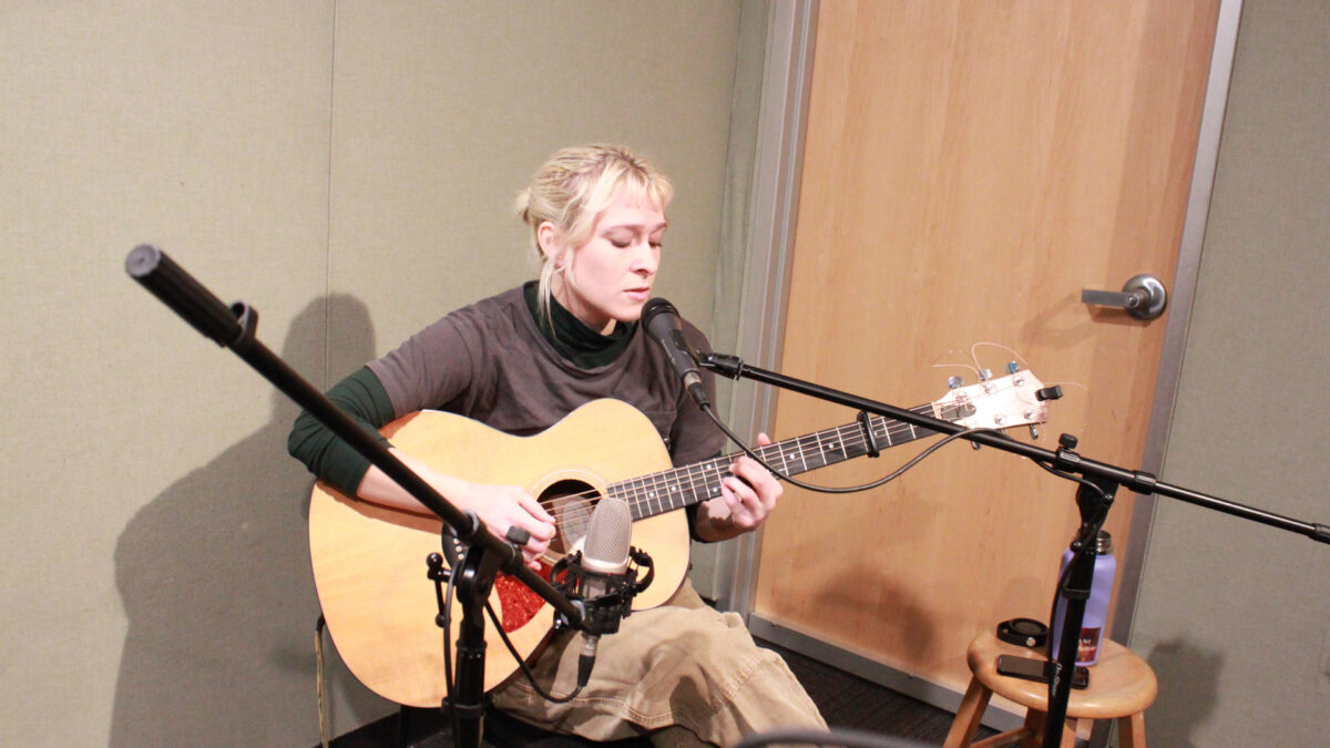 An adult sitting while playing acoustic guitar and singing into a microphone in a radio studio