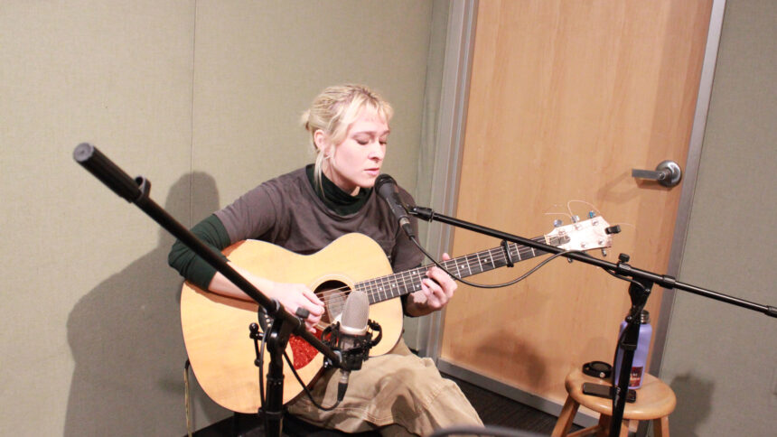 An adult sitting while playing acoustic guitar and singing into a microphone in a radio studio