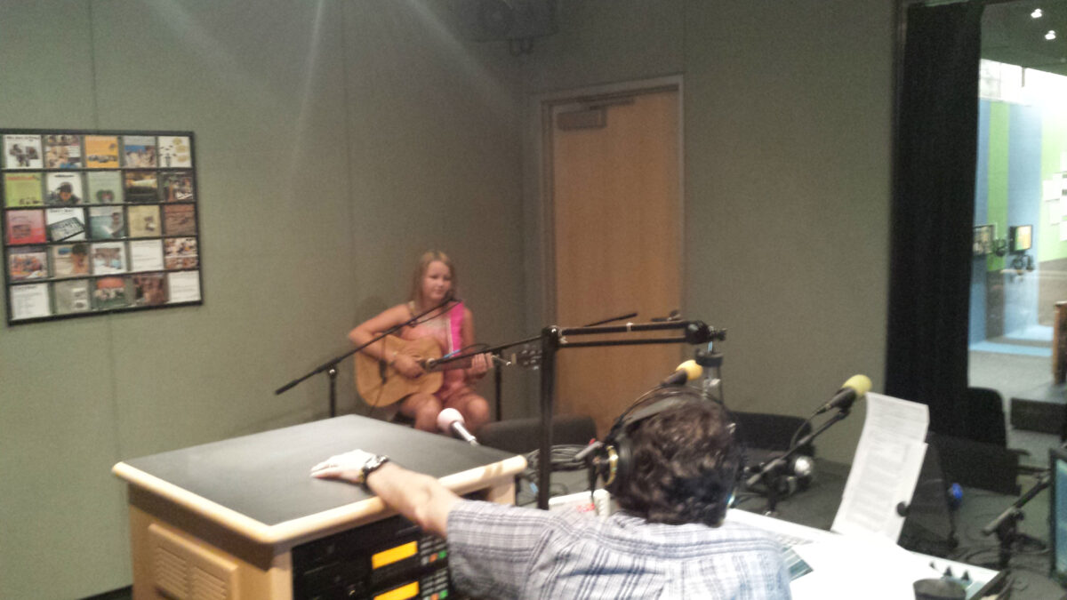 A youth holding an acoustic guitar and sitting behind a microphone being interviewed by an adult wearing headphones sitting behind a microphone and a mixer in a radio studio