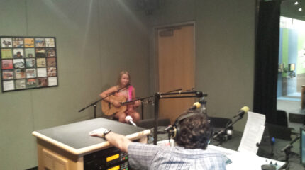 A youth holding an acoustic guitar and sitting behind a microphone being interviewed by an adult wearing headphones sitting behind a microphone and a mixer in a radio studio