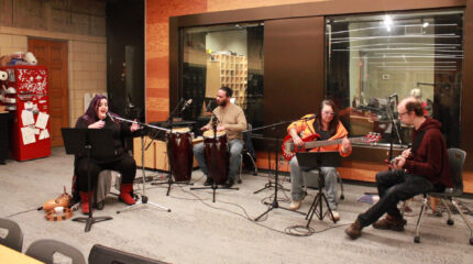 An adult singing into a microphone, an adult playing congas and sitting behind a microphone, an adult sitting behind a microphone playing an electric bass, and an adult sitting behind a microphone playing an electric guitar