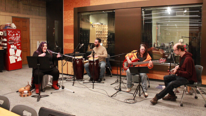 An adult singing into a microphone, an adult playing congas and sitting behind a microphone, an adult sitting behind a microphone playing an electric bass, and an adult sitting behind a microphone playing an electric guitar