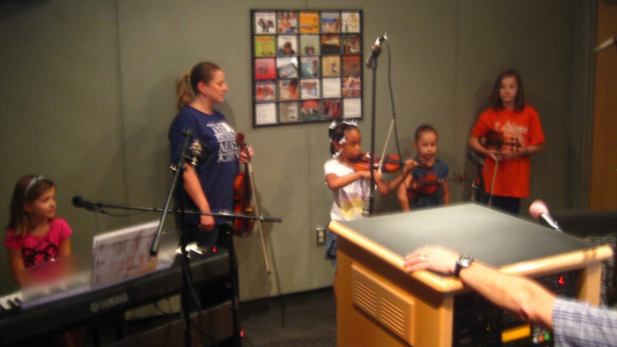 A youth sitting behind a piano keyboard and a microphone and an adult holding a violin looking down at three youth holding violins standing behind a microphone in a radio studio