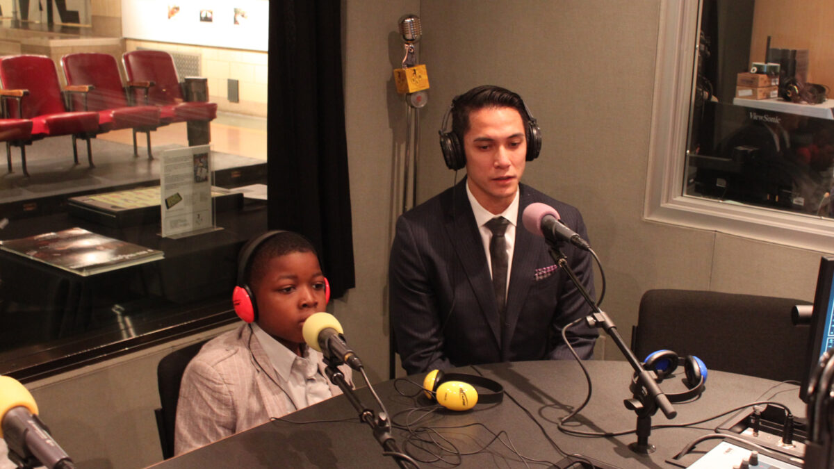An adult and a youth wearing headphones speaking into microphones in a radio studio