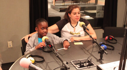 A youth and an adult sitting behind microphones and headphones in a radio studio