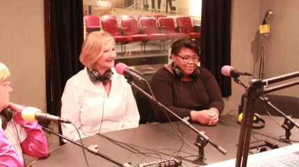 Two adults and a teen wearing headphones around their necks speaking into microphones in a radio studio