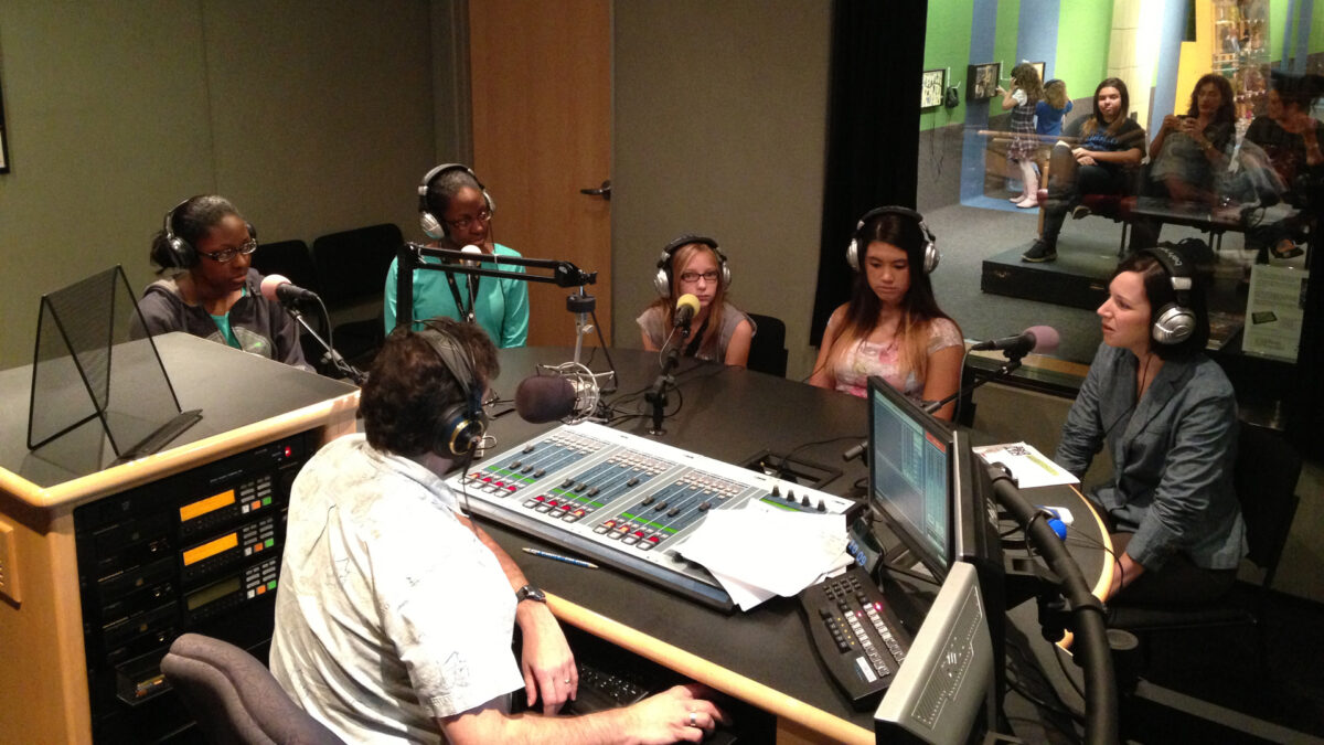 Four youth and one adult wearing headphones sitting behind microphones being interviewed by an adult wearing headphones sitting behind a microphone and a mixing board in a radio studio