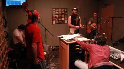An adult sitting at a drum set, an adult holding an upright bass, an adult playing a banjo and singing into a microphone, and an adult holding an acoustic guitar and standing behind a microphone, all being interviewed by an adult wearing headphones sitting behind a mixer in a radio studio