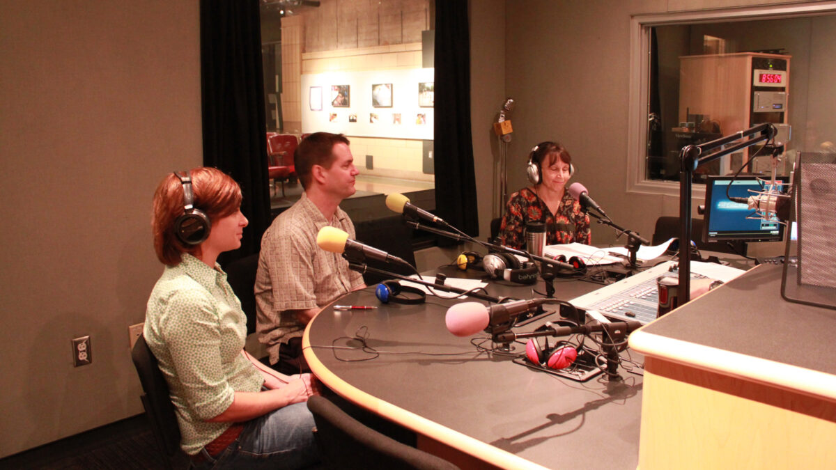 Three adults, two wearing headphones, speaking into microphones in a radio studio.