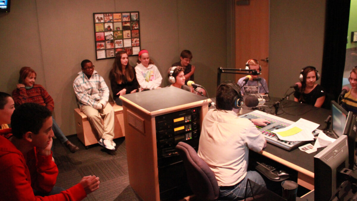 Seven youth on the edge of the room looking at four youth wearing headphones and speaking into microphones being interviewed by an adult wearing headphones sitting behind a microphone and a mixer in a radio studio