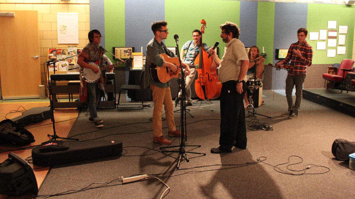 An adult holding a banjo, an adult sitting at a piano keyboard, an adult leaning on an upright bass, an adult sitting behind a snare drum, an adult holding a violin, and an adult holding a microphone interviewing an adult holding an acoustic guitar.