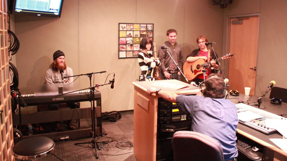 An adult sitting at a piano keyboard, an adult standing with their arms folded, and two adults holding acoustic guitars, all behind microphones, being interviewed by an adult wearing headphones sitting behind a mixer in a radio studio