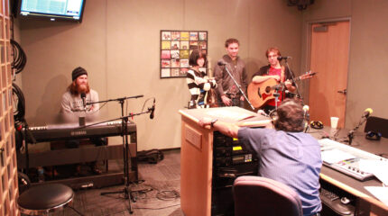 An adult sitting at a piano keyboard, an adult standing with their arms folded, and two adults holding acoustic guitars, all behind microphones, being interviewed by an adult wearing headphones sitting behind a mixer in a radio studio