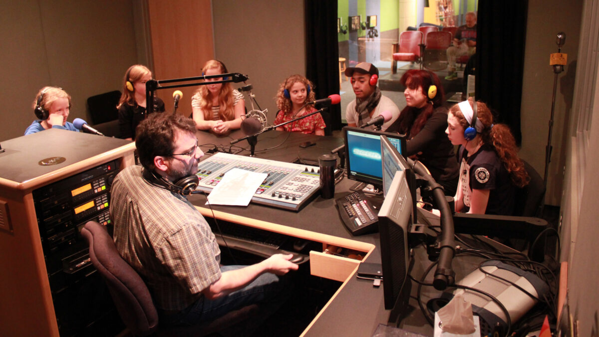 Seven youth wearing headphones sitting behind microphones being interviewed by an adult wearing headphones and sitting behind a microphone and a mixing board in a radio studio