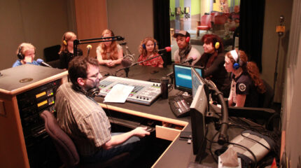 Seven youth wearing headphones sitting behind microphones being interviewed by an adult wearing headphones and sitting behind a microphone and a mixing board in a radio studio