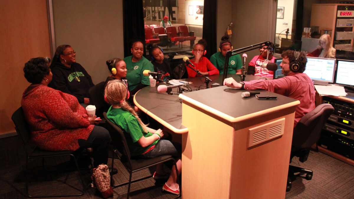 Seven youth wearing headphones and two adults, all sitting behind microphones being interviewed by an adult wearing headphones sitting behind a microphone in a radio studio