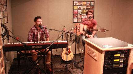 An adult sitting behind a piano keyboard and an adult sitting while holding an acoustic guitar, both behind microphones in a radio studio