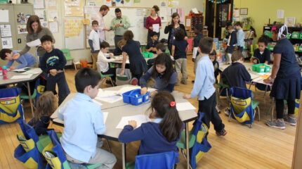 Eighteen youth, eleven sitting at classroom desks, seven standing around the desks, and five adults overseeing while standing in front of a whiteboard.