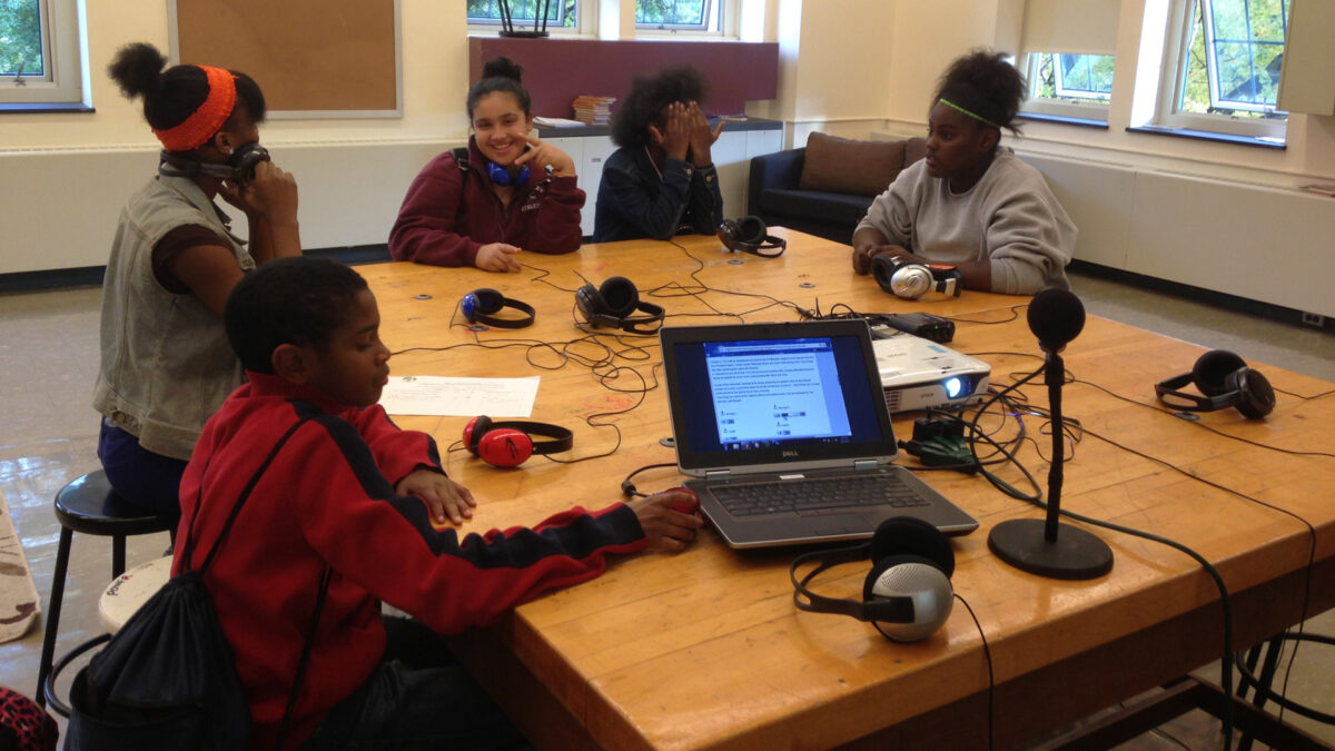 Five youth sitting around pairs of headphones connected to a laptop computer and a microphone on a wooden table in a classroom