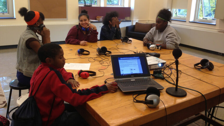 Five youth sitting around pairs of headphones connected to a laptop computer and a microphone on a wooden table in a classroom