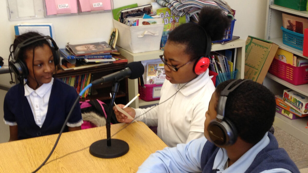 Three youth wearing headphones sitting around a microphone on a classroom desk