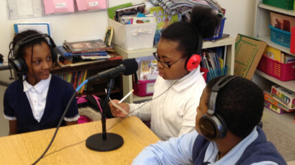 Three youth wearing headphones sitting around a microphone on a classroom desk