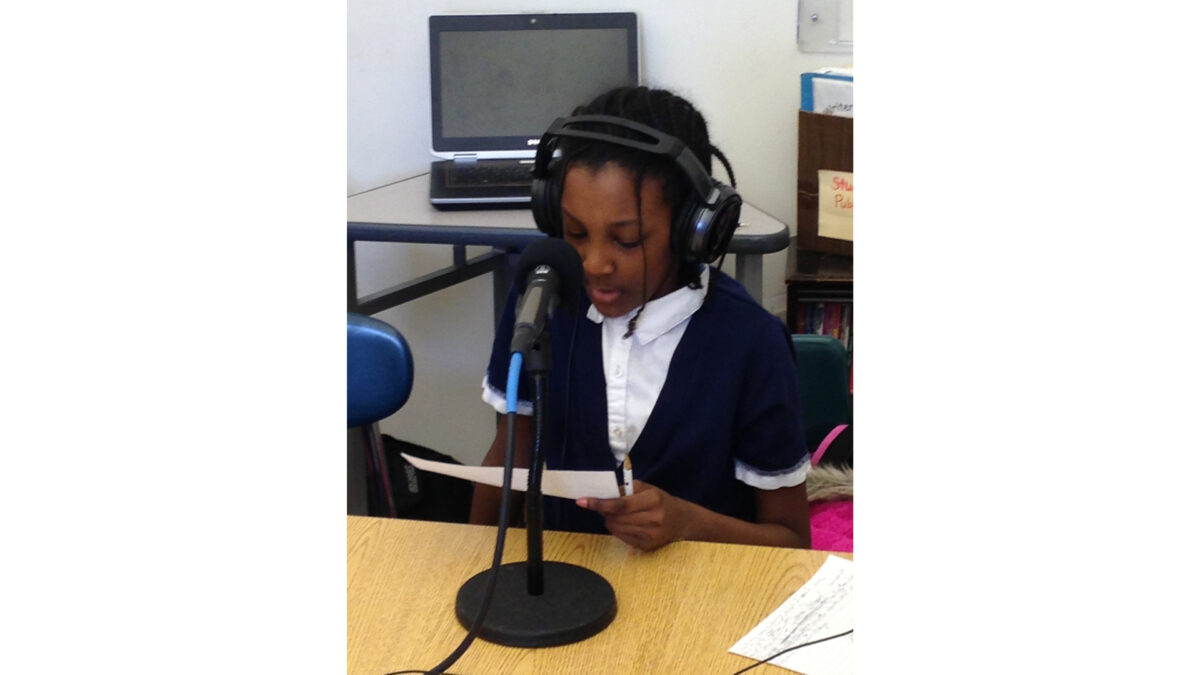 A youth wearing headphones sitting behind a microphone on a classroom desk and reading a piece of paper