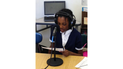 A youth wearing headphones sitting behind a microphone on a classroom desk and reading a piece of paper
