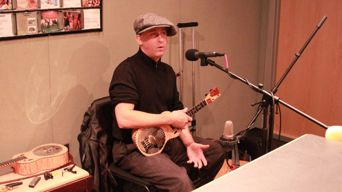An adult sitting while holding a ukulele and speaking into a microphone in a radio studio