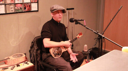 An adult sitting while holding a ukulele and speaking into a microphone in a radio studio
