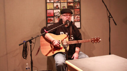 An adult sitting while holding an acoustic guitar and speaking into a microphone in a radio studio