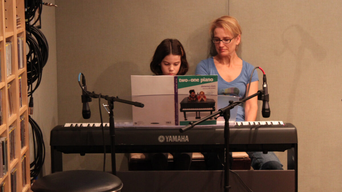 An adult looking at a youth playing a piano keyboard in a radio studio